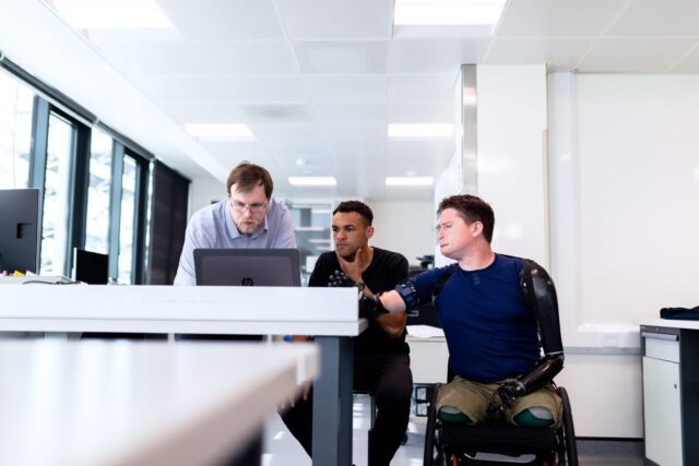 three men, one using a wheelchair, look at a computer screen